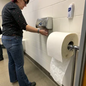 A woman reaches for a paper towel from a dispenser in a restroom, with a roll of paper towels visible.